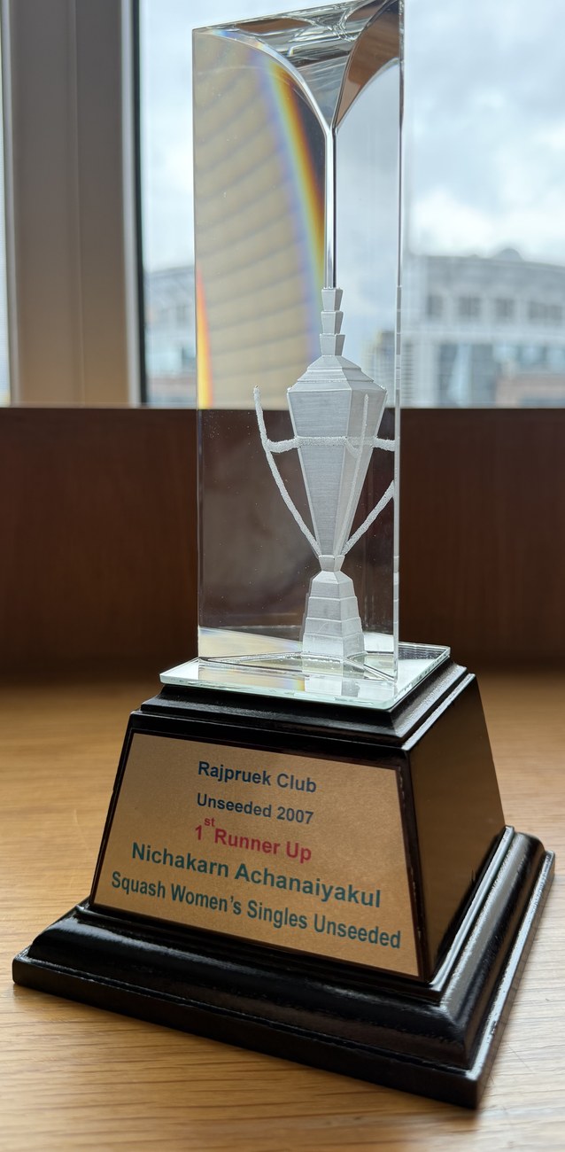 A clear glass squash trophy on a black wooden base, engraved with a cup and a racket, its brass plaque reading Rajpruek Club, Unseeded 2007, 1st Runner Up, Nichakarn Achanaiyakul, Squash Women’s Singles Unseeded
