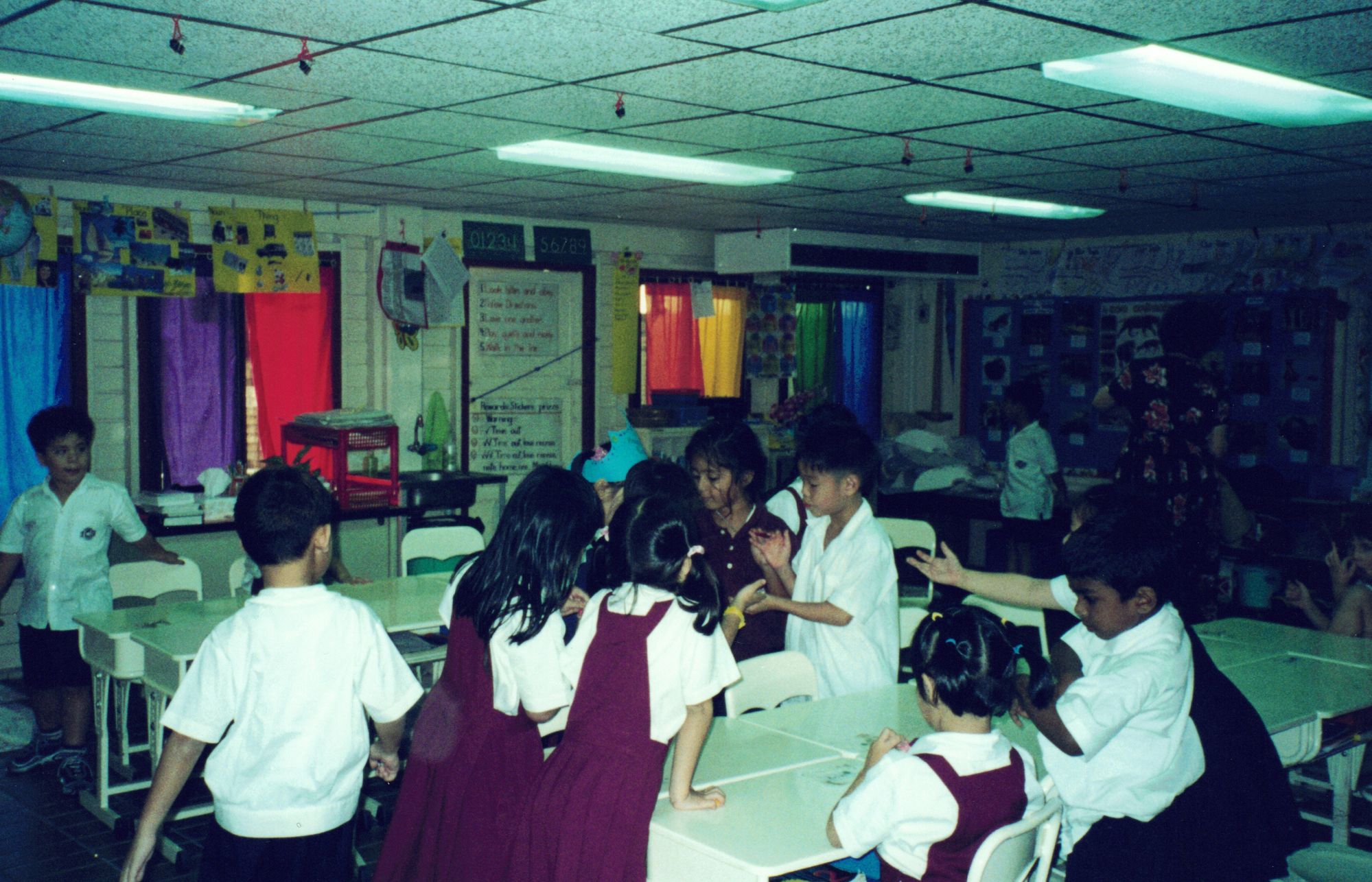 Classmates in maroon pinafores gathered around tables in a classroom