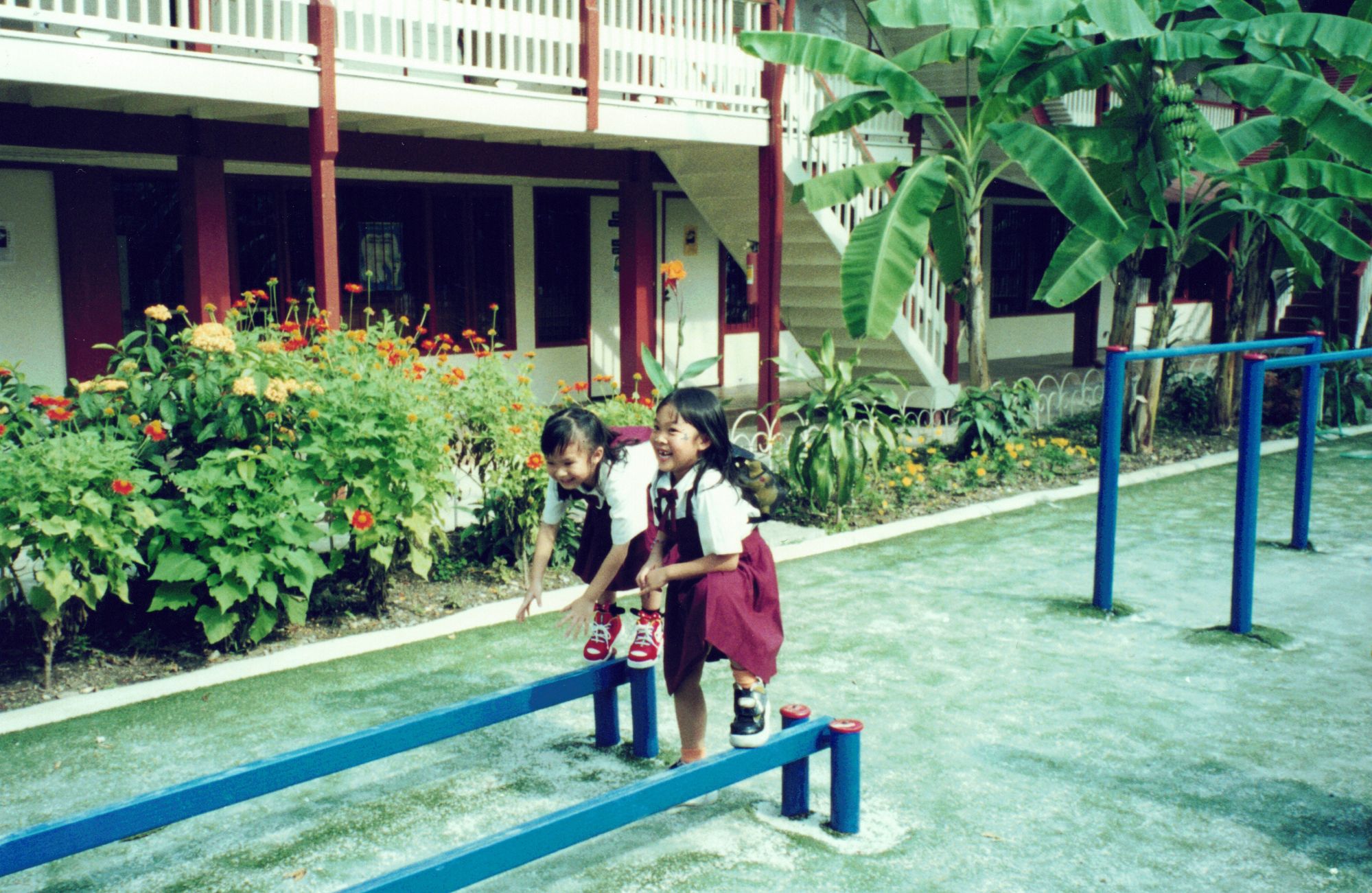 Two girls in school uniform balancing along a low beam in the school garden