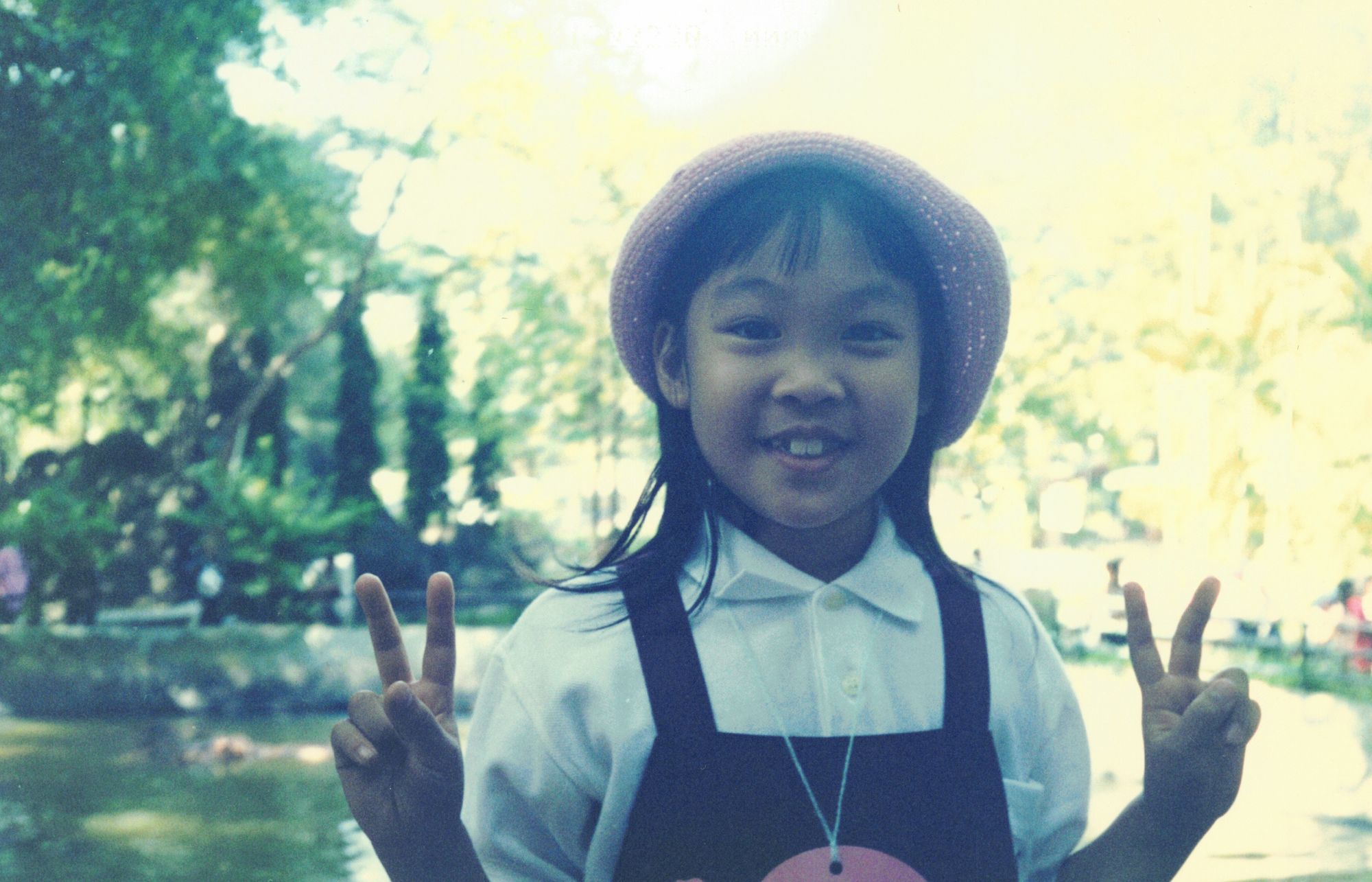 Me in a woven hat by a pond on a school trip, making peace signs at the camera