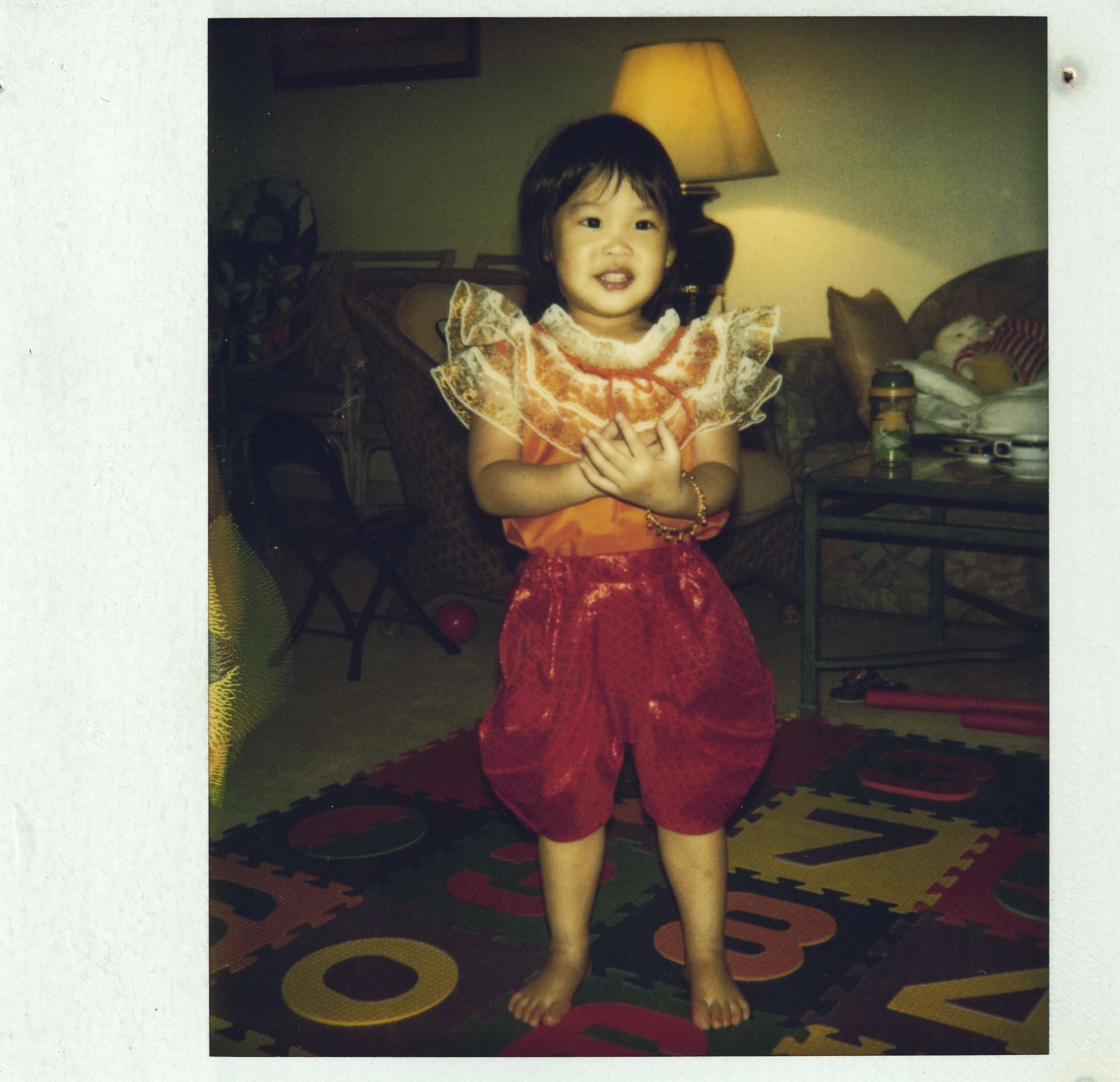 Me as a toddler in a red Thai costume, standing on a play mat
