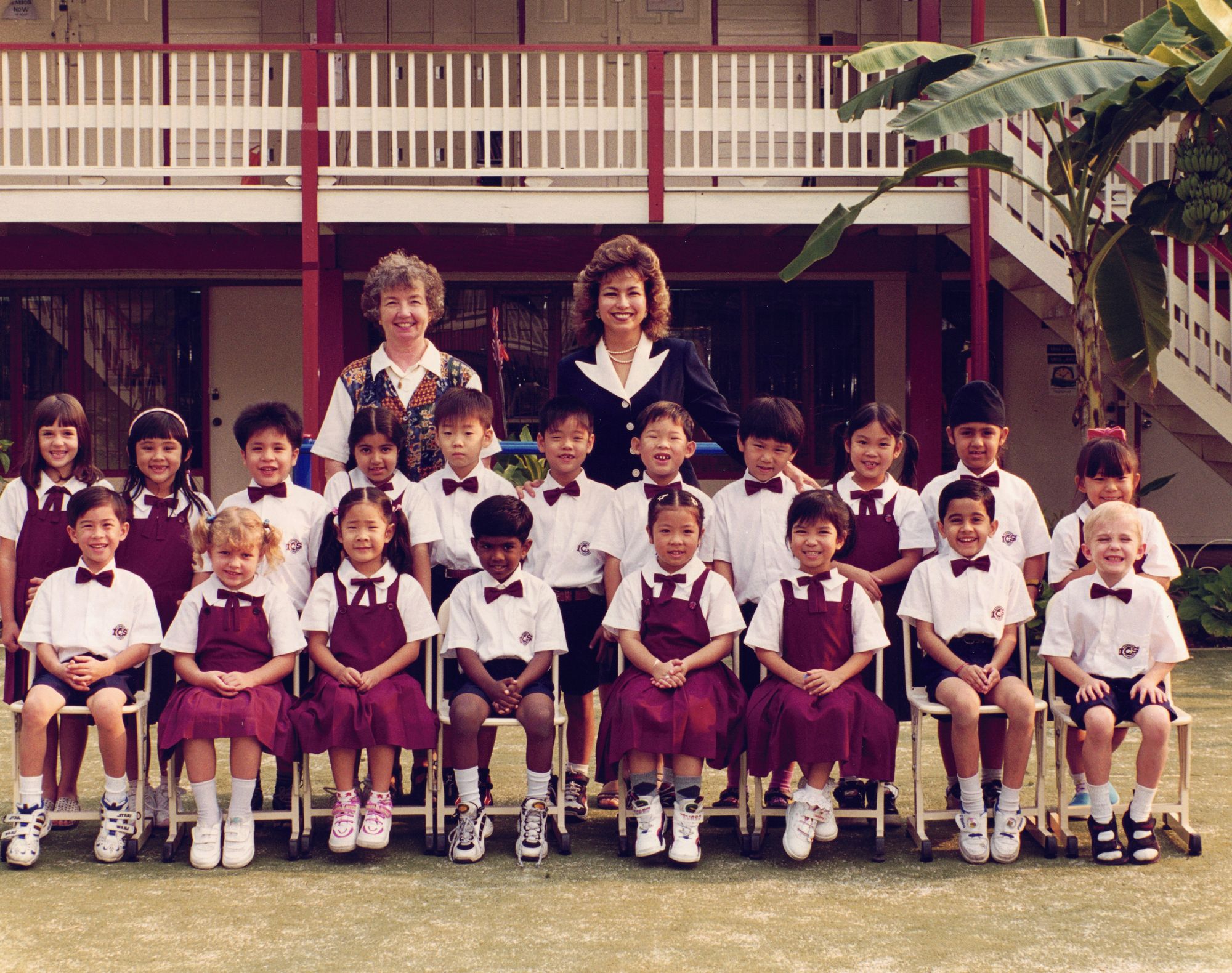 My ICS kindergarten class photograph — children in white shirts and maroon pinafores seated on the school steps, two teachers standing at the back