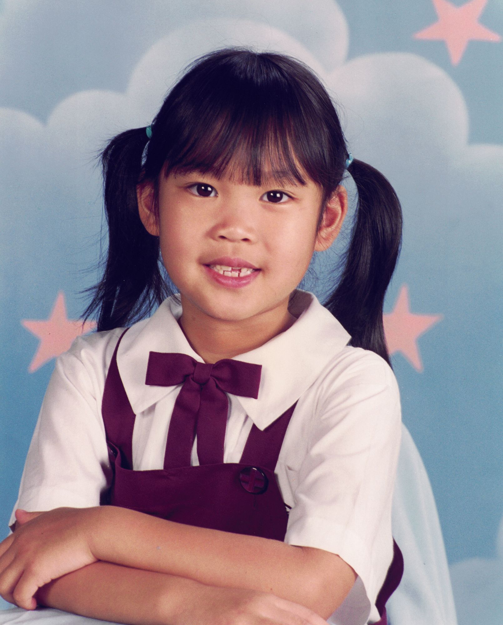 My ICS kindergarten studio portrait — pigtails, white shirt, maroon pinafore, against a sky-and-stars backdrop