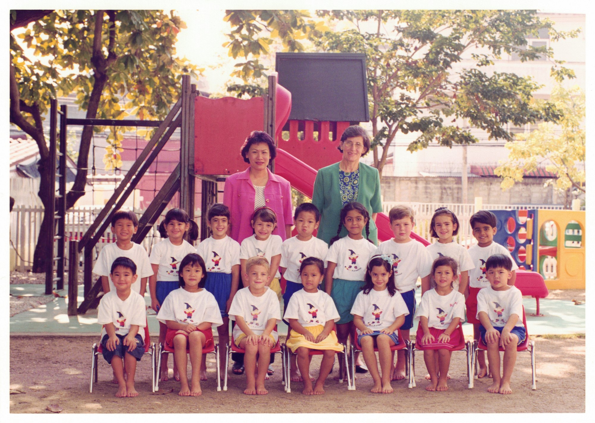 My Noddy preschool class in the playground — children in white shirts seated in rows, two teachers standing behind